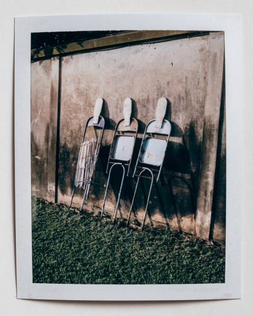 Three metal folding chairs stand against a concrete wall outdoors on a sunny day, casting shadows on the wall and green grass below.