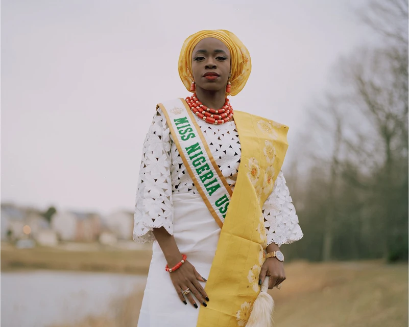 Portrait of a smiling woman in traditional Nigerian formalwear posing outdoors with a lake in the background on an overcast day.