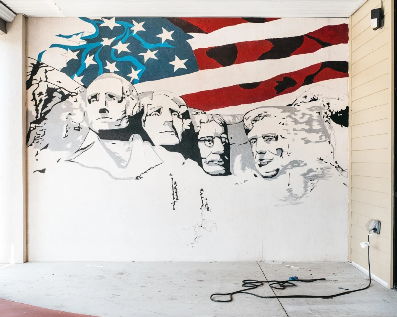 Painted mural on a wall showing Mount Rushmore overlaid with an American flag, rendered in shades of gray, blue, and red.