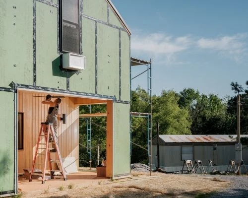 Construction worker on a ladder repairing the exterior of a house with green siding, with trees and blue sky in the background.