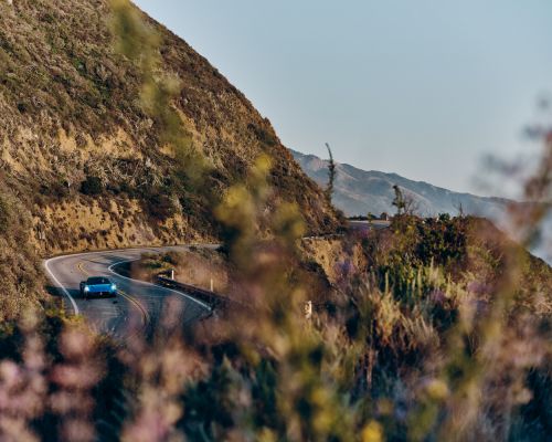 Blue convertible car driving on a winding coastal road, backed by green mountain slopes and a distant mountain range, bright daylight.