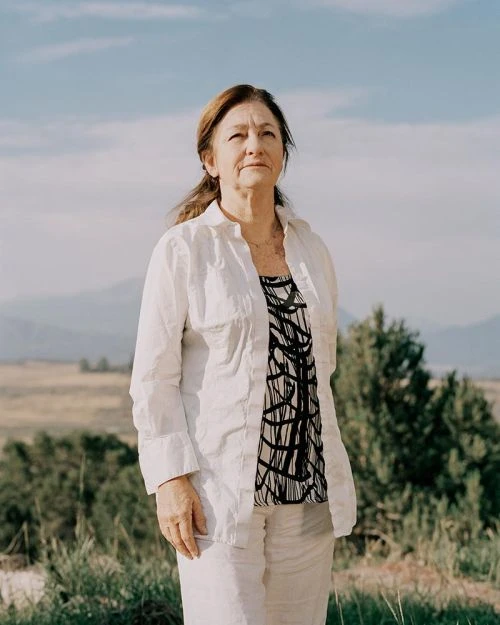 Portrait of an older woman in a white linen outfit, standing outdoors against a scenic background of mountains and blue sky.