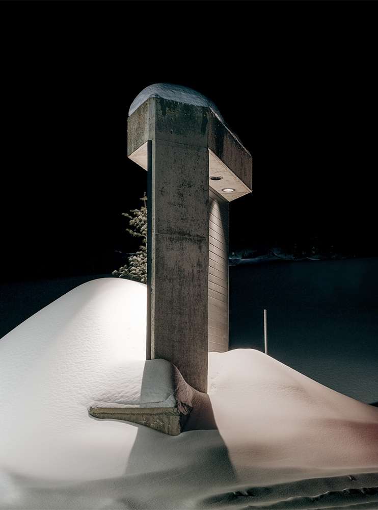 Monochrome photograph of a concrete architectural structure covered in snow at night with a black background and a snow covered landscape.