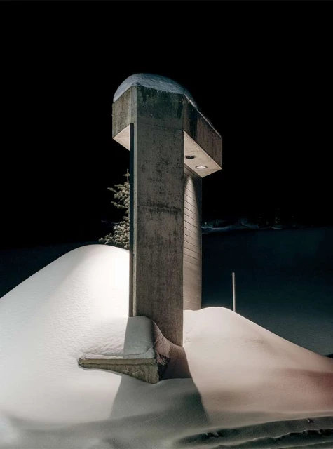 Monochrome photograph of a concrete architectural structure covered in snow at night with a black background and a snow covered landscape.