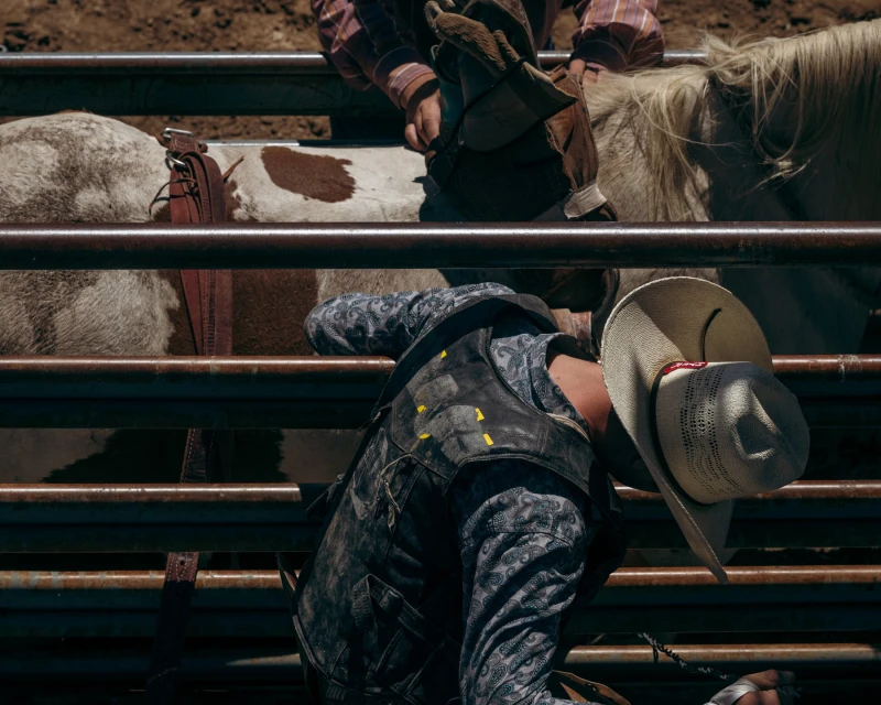 Rodeo athlete on bucking bronco in outdoor arena; the cowboy wears a hat and vest as a second cowboy looks on from behind.