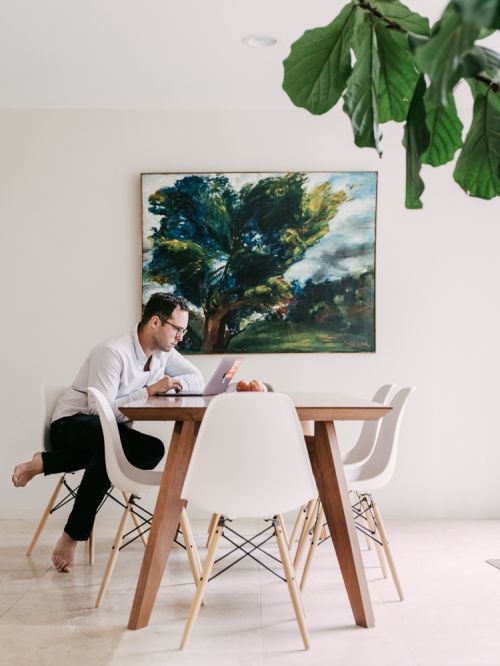 Adult male working on a laptop at a wooden table with white chairs in an indoor setting, with a tree painting on the wall.