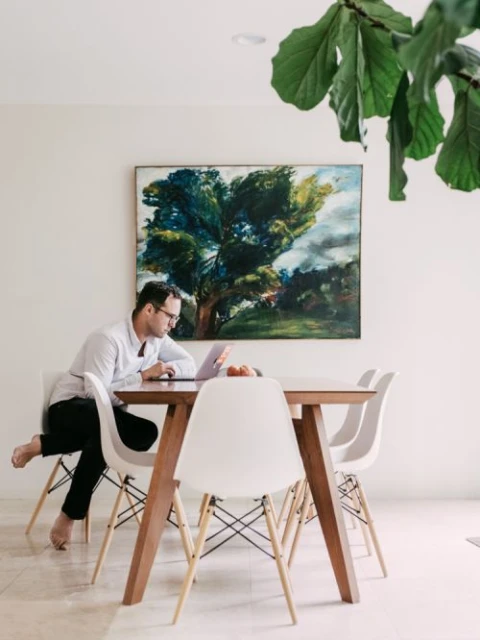 Adult male working on a laptop at a wooden table with white chairs in an indoor setting, with a tree painting on the wall.