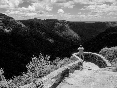 Monochrome landscape view of person photographing mountain valley from overlook with stone wall and cloudy sky in black and white.