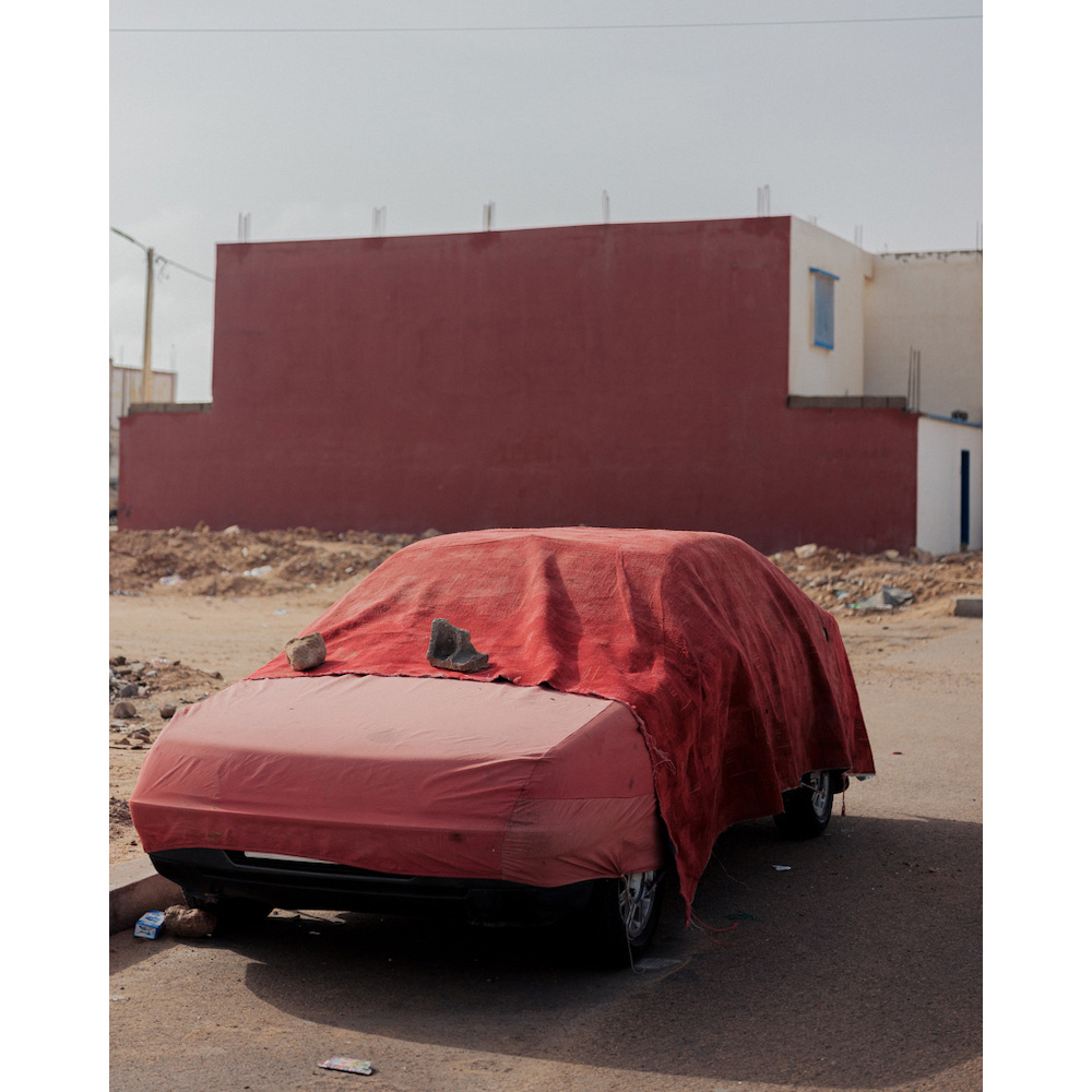 Front view of a car covered with a large red cloth, parked on an asphalt road, with a large red building in the background on a sunny day.