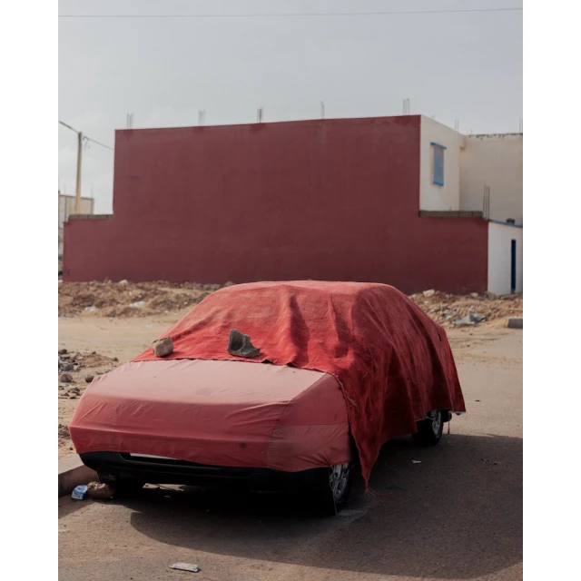Front view of a car covered with a large red cloth, parked on an asphalt road, with a large red building in the background on a sunny day.