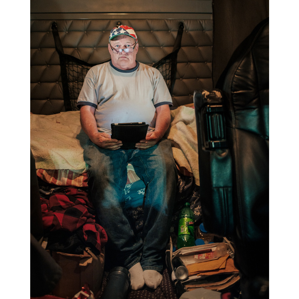 Truck driver sits in his cab, using a tablet, surrounded by personal belongings, showcasing the interior of a long-haul vehicle.