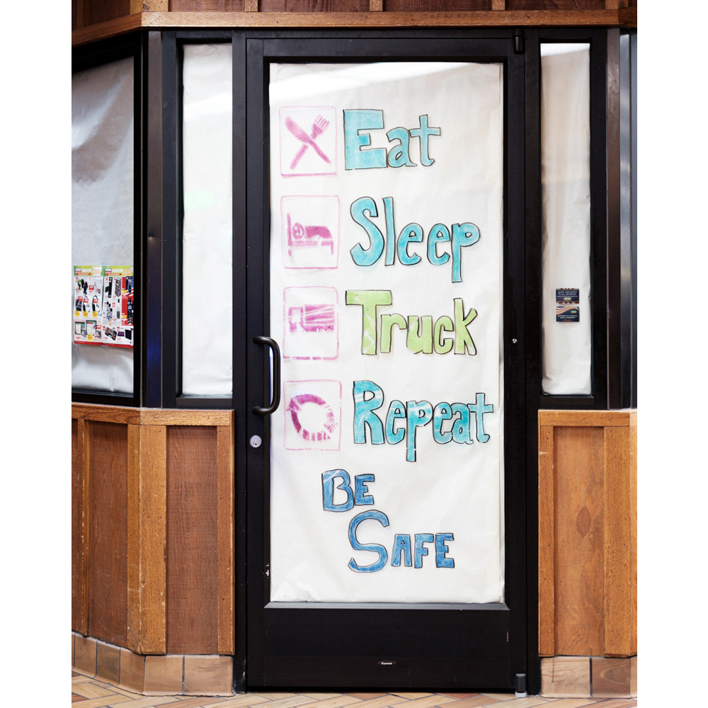 Black-framed glass door with colorful handwritten message inside a building with wood paneling and tile floor, promoting truck stop services.