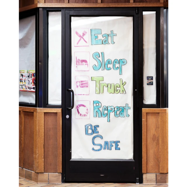 Black-framed glass door with colorful handwritten message inside a building with wood paneling and tile floor, promoting truck stop services.
