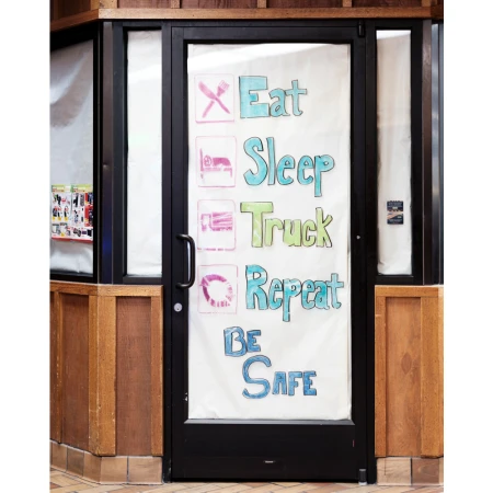 Black-framed glass door with colorful handwritten message inside a building with wood paneling and tile floor, promoting truck stop services.