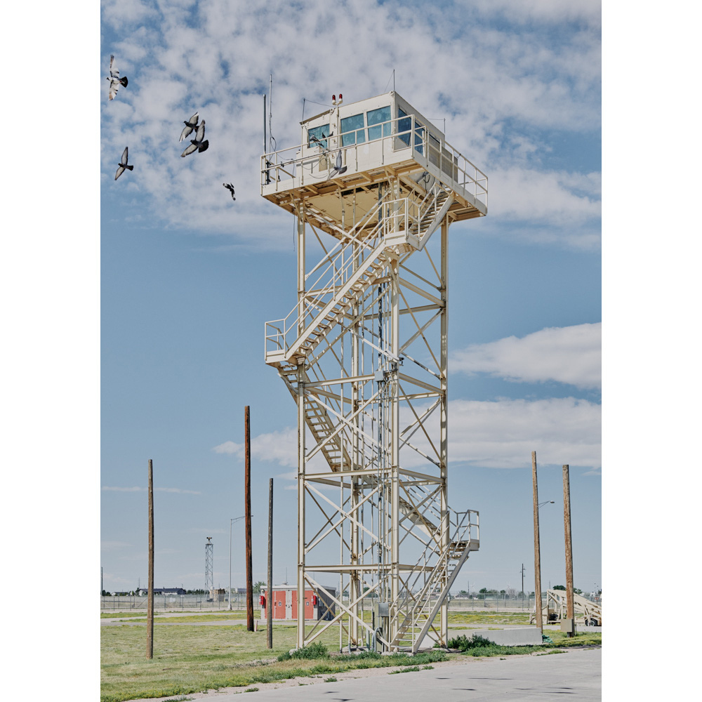 Tall white metal control tower with exterior stairs and a glass-enclosed top, with flying birds against a blue sky and a flat, grassy landscape.