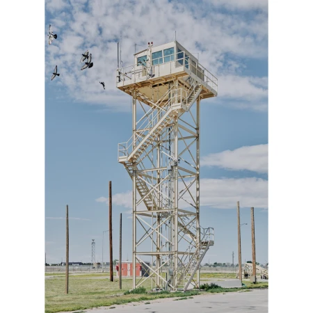 Tall white metal control tower with exterior stairs and a glass-enclosed top, with flying birds against a blue sky and a flat, grassy landscape.