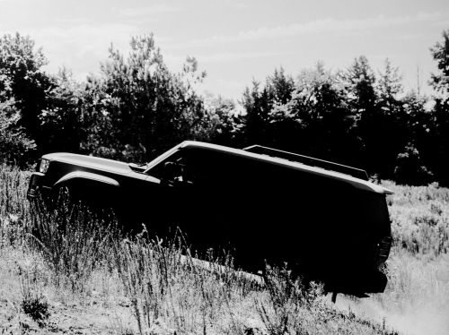Monochrome photograph of a dark SUV on a grassy hill against a backdrop of trees and a cloudy sky; high contrast and dramatic lighting.
