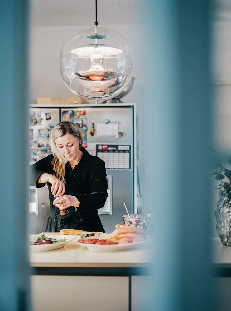 Woman in black shirt prepares food in a kitchen with overhead lighting. Ingredients sit on the counter. Clear vertical foreground blur.