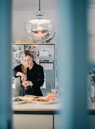 Woman in black shirt prepares food in a kitchen with overhead lighting. Ingredients sit on the counter. Clear vertical foreground blur.