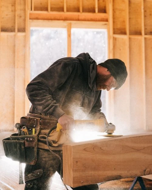Carpenter sands wood with a power tool indoors; wood structure and bright light highlight the construction scene and activity.