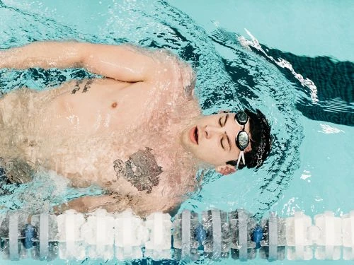 A shirtless man wearing goggles swims the backstroke in an indoor swimming pool with clear blue water and a lane divider.