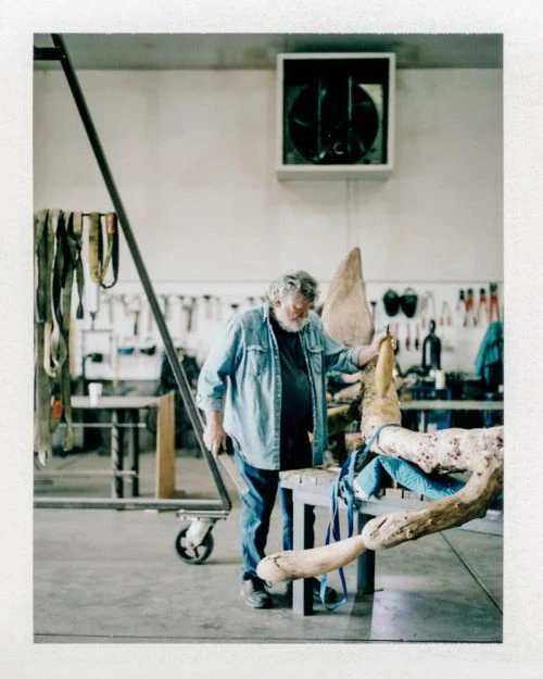 An elderly man works on a light-colored wooden sculpture in a workshop with tools and equipment visible in the background.