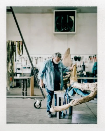 An elderly man works on a light-colored wooden sculpture in a workshop with tools and equipment visible in the background.