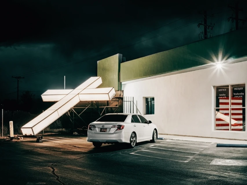 Night scene of a white car parked near a building with a large illuminated cross, under a dark, cloudy sky in an outdoor setting.