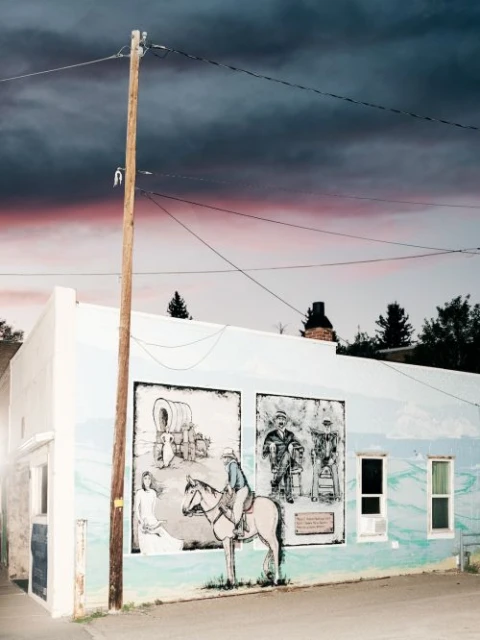 Outdoor mural depicting historical figures and scenes painted on the side of a white building with a telephone pole and cloudy sky.