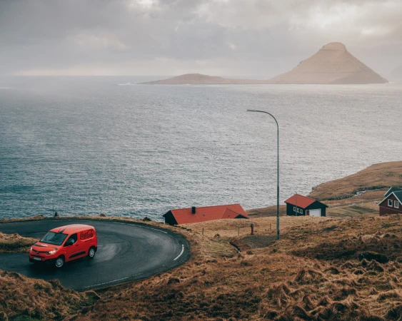Red delivery van on winding road along the coast in the Faroe Islands with mountains and ocean in background on an overcast day.