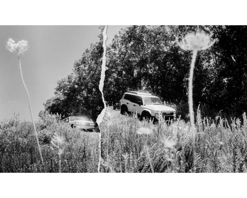 Monochrome landscape with a vintage car and a white SUV parked on a grassy hill under a clear sky, creating a nostalgic, rural scene.