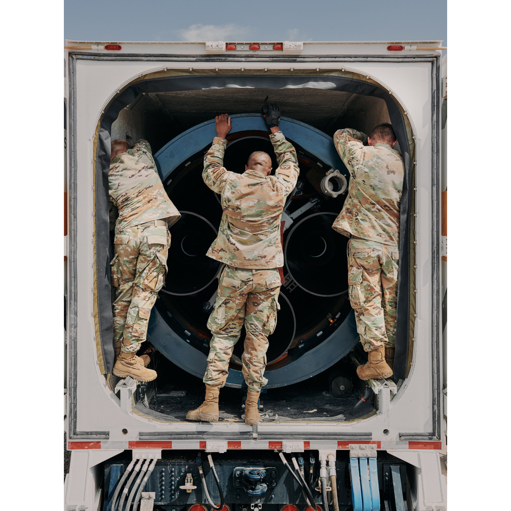 Three uniformed service members load a large cylindrical piece of equipment into a white truck, showcasing a scene of logistics.