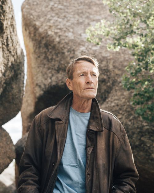 Portrait of a mature man in a brown leather jacket outdoors with large boulders and foliage. Man gazes to the right of the frame.