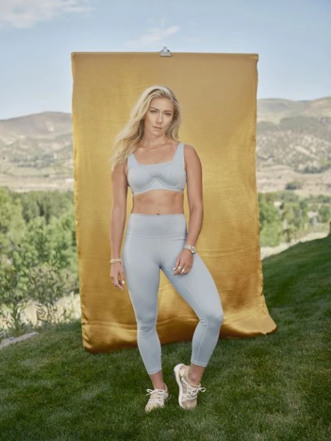 Athletic woman in fitness attire poses outdoors with a gold backdrop, set against a backdrop of green grass and a mountain view.