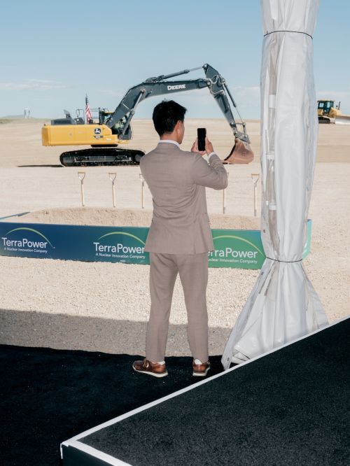 Adult male takes picture of a yellow excavator with a smartphone in a sunny, outdoor construction environment during daylight.