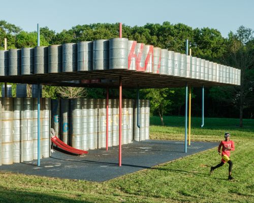Athletic man in sportswear runs past an outdoor metal-barrel art installation in the green grassy yard on a sunny day.