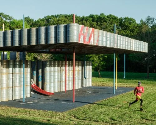 Athletic man in sportswear runs past an outdoor metal-barrel art installation in the green grassy yard on a sunny day.
