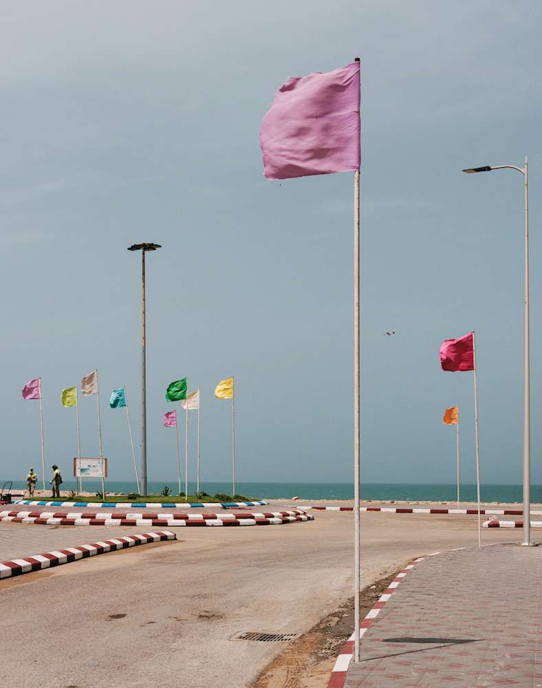 Colorful flags on poles line a coastal road with ocean backdrop under a blue sky, creating a vibrant and inviting scene.