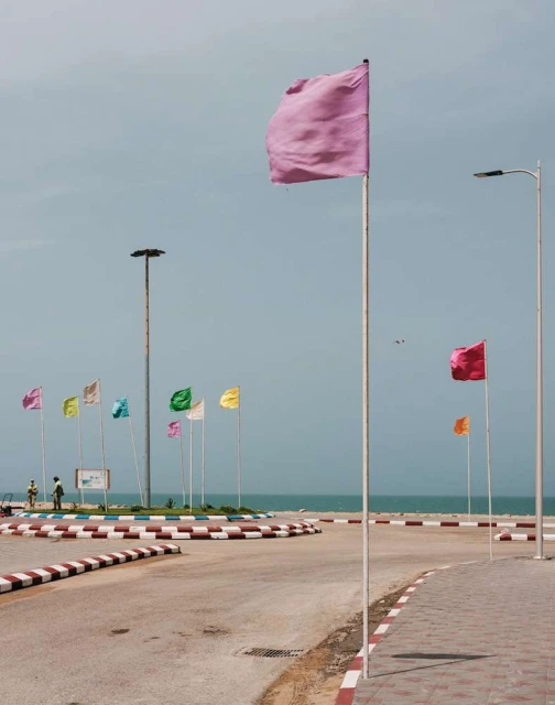 Colorful flags on poles line a coastal road with ocean backdrop under a blue sky, creating a vibrant and inviting scene.