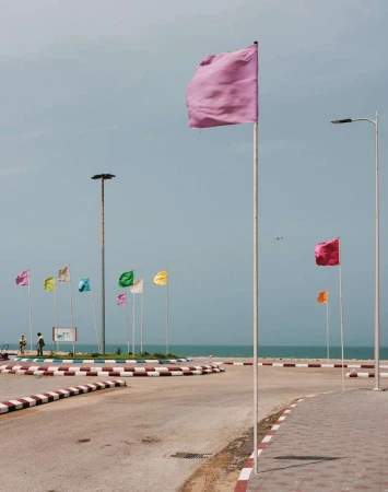 Colorful flags on poles line a coastal road with ocean backdrop under a blue sky, creating a vibrant and inviting scene.