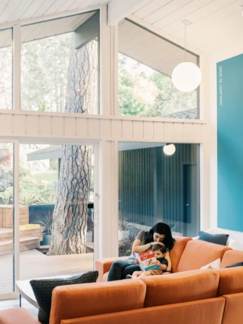Mother and child read a book together on an orange sofa in a sun-lit modern living room with large windows and outdoor views.