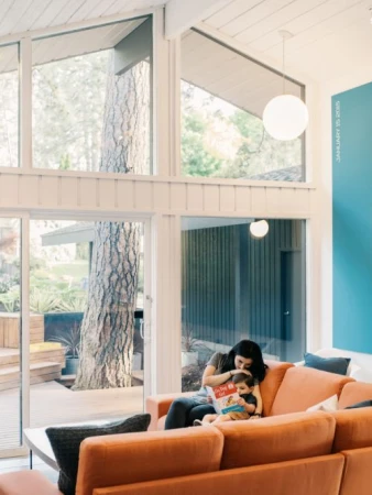 Mother and child read a book together on an orange sofa in a sun-lit modern living room with large windows and outdoor views.