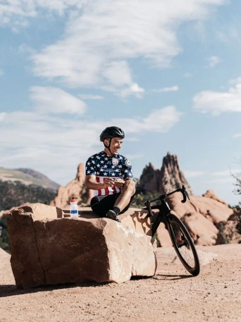 A man in cycling gear sits on a rock with his road bike, water bottle, and snack in a sunny, mountainous outdoor setting.