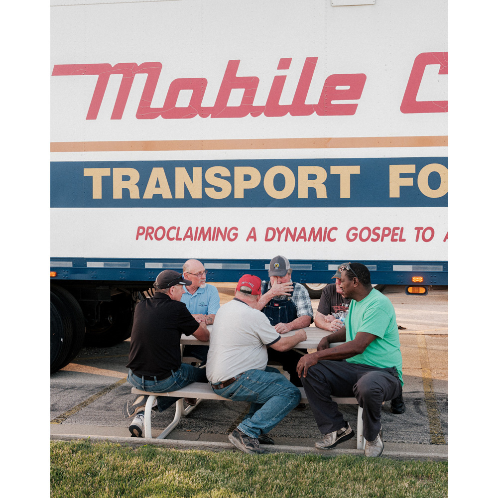 Six men gather around a picnic table outside a Mobile Care Transport vehicle for a meeting, sharing a meal, or break.