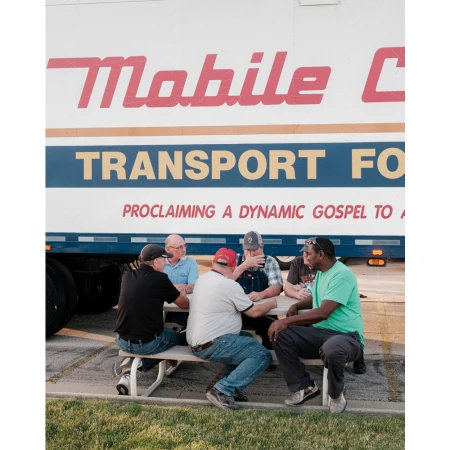 Six men gather around a picnic table outside a Mobile Care Transport vehicle for a meeting, sharing a meal, or break.