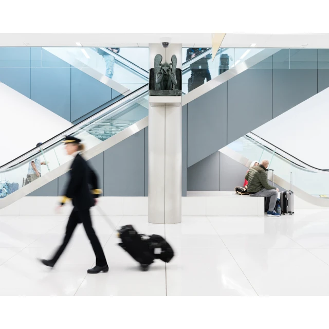 Motion blur of pilot pulling rolling suitcase through brightly lit airport, architectural steel and glass design with an escalator and seating.