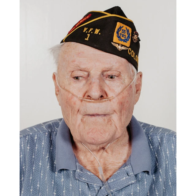 Elderly man in a military hat with insignias, wearing a blue shirt and oxygen tubes in a studio setting, captured in a medium shot.