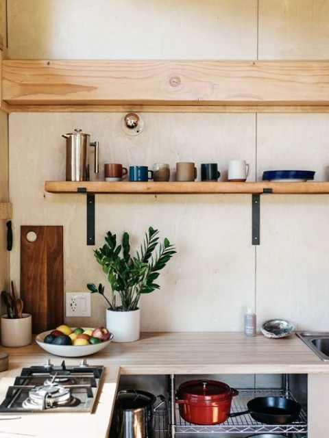Bright, modern kitchen with wood cabinets, a plant, colorful mugs on a shelf, plus cooking equipment, bowls of fruit, and cookware.