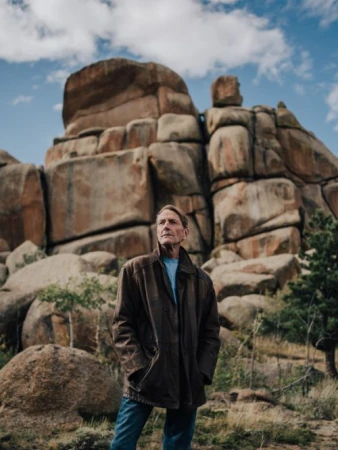Elderly man wearing a brown leather jacket and blue jeans standing before massive rock formations