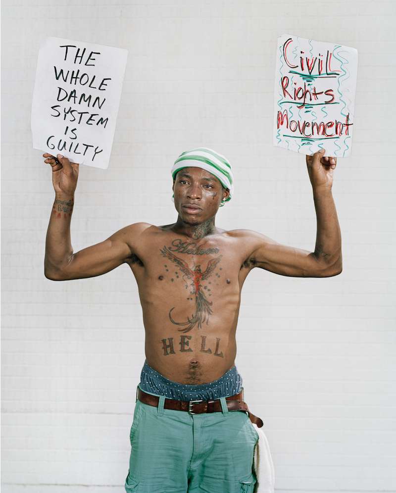 Shirtless man holds protest signs with text against a white brick background, wearing a cap and casual shorts with visible tattoos. 